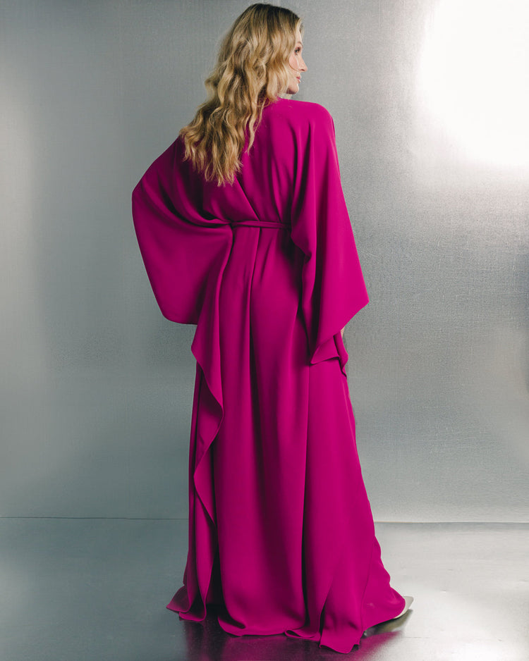 Woman in elegant fuchsia maxi dress with flowing sleeves, standing in minimal studio setting