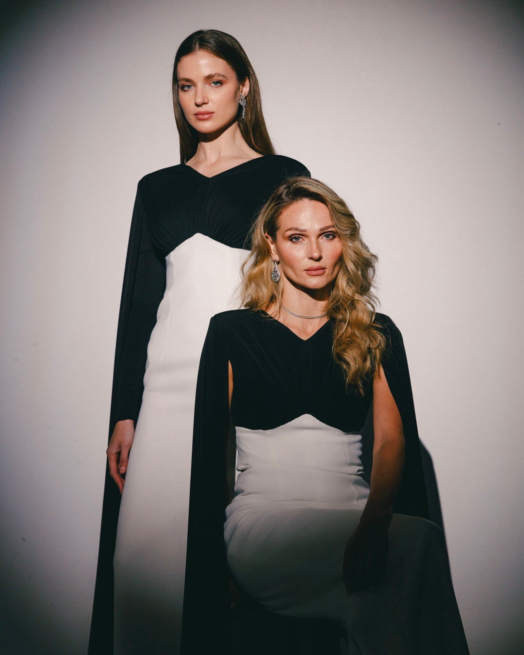 Two women in black and white evening gowns with cape sleeves, posing against a neutral background