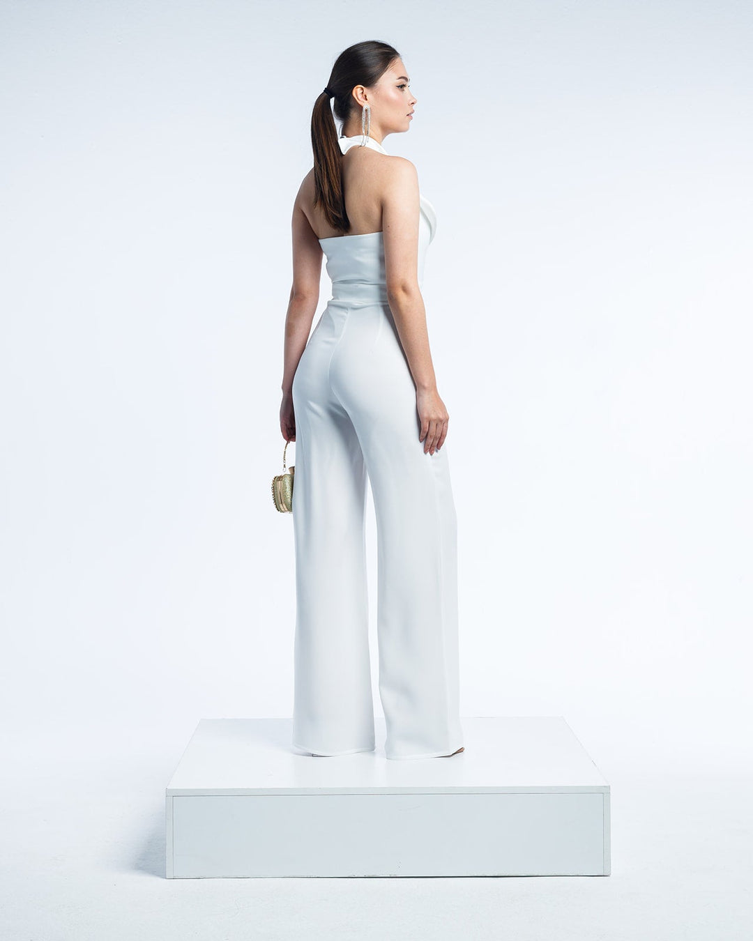 Woman wearing a white halter-neck jumpsuit, wide-leg pants, holding a gold clutch