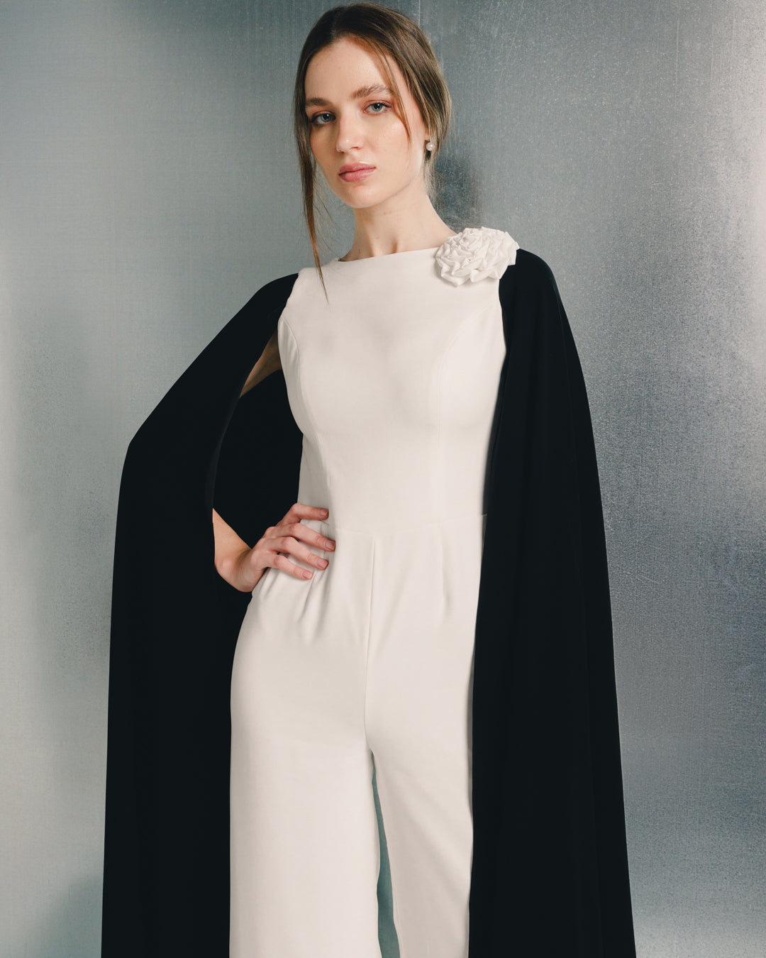 Woman in elegant white jumpsuit with floral shoulder detail and black cape, fashion photoshoot