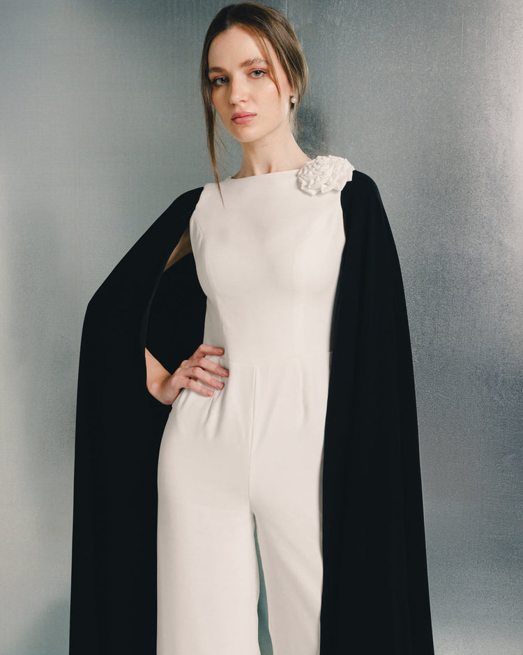 Woman in elegant white jumpsuit with floral shoulder detail and black cape, fashion photoshoot