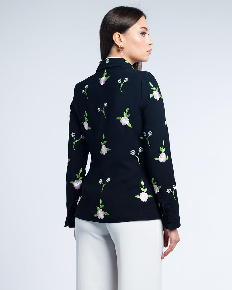 Woman wearing black floral embroidered blazer with white trousers, back view