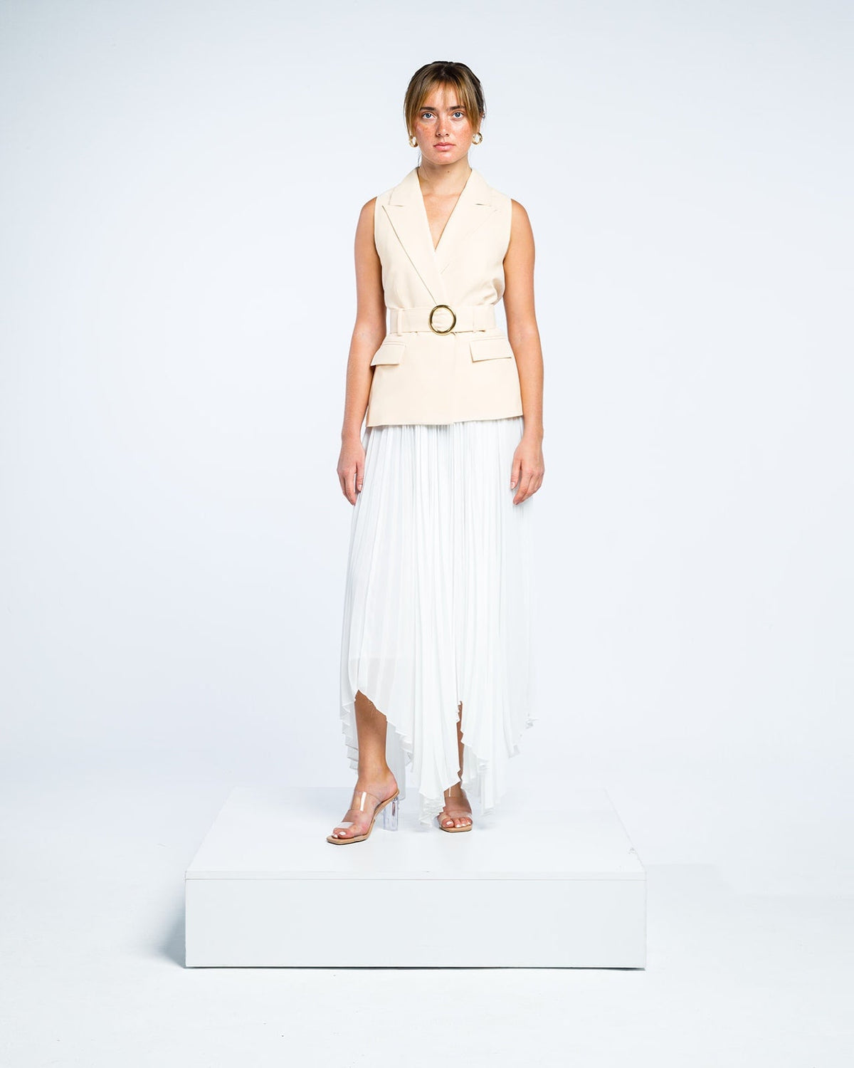 Woman wearing a sleeveless beige belted vest and white pleated midi skirt, standing on a white platform.