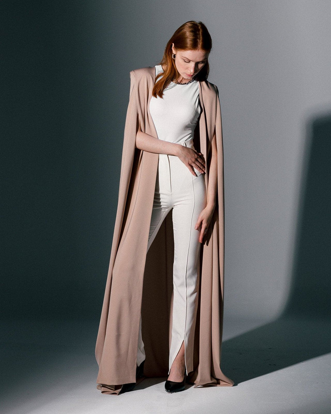 Woman in ivory jumpsuit with long beige crepe cape, elegant modern fashion style