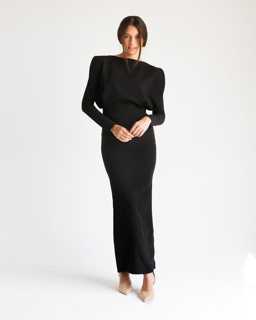 Woman in a long black elegant dress with structured shoulders, standing in a minimal studio
