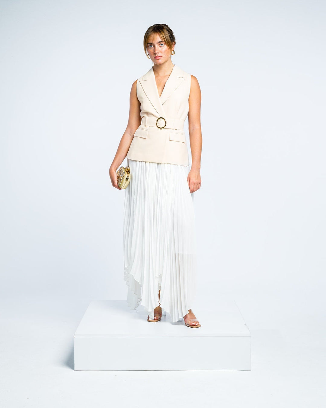 Woman in a sleeveless beige belted blazer and white pleated maxi skirt holding clutch