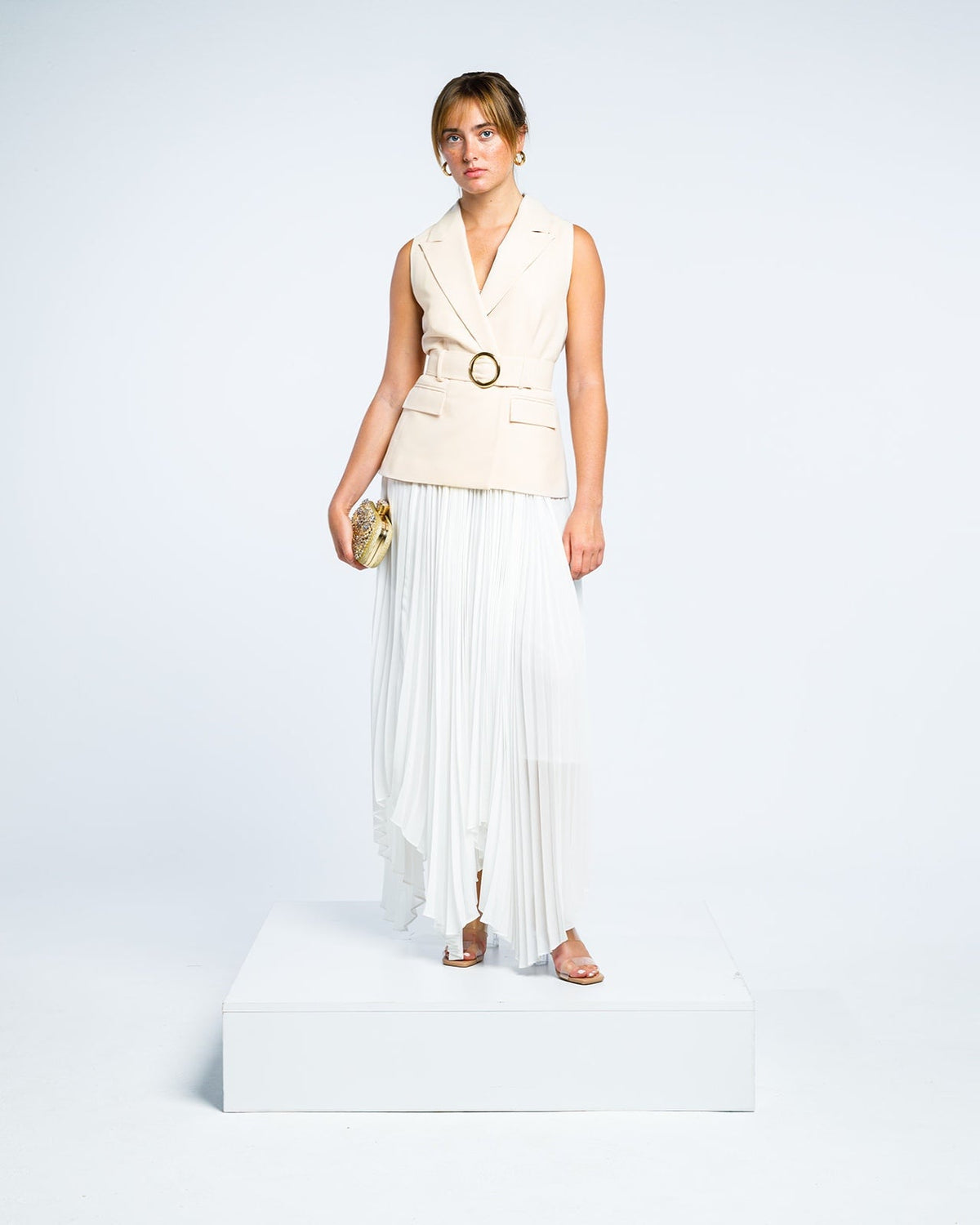 Woman in a sleeveless beige belted blazer and white pleated maxi skirt holding clutch