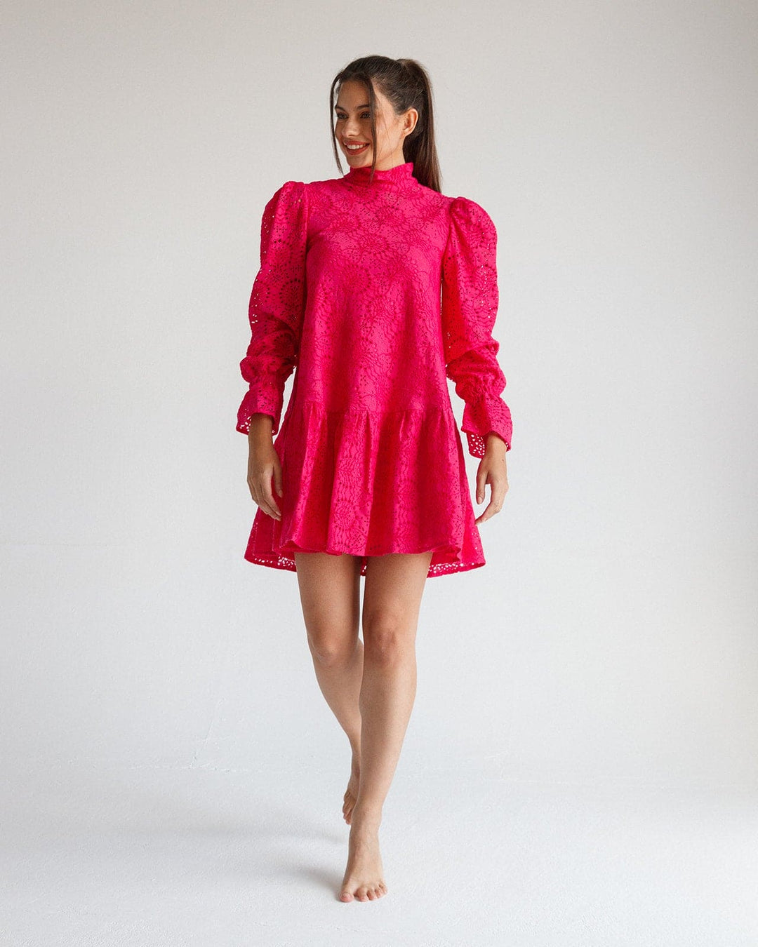 Woman in a bright fuchsia lace mini dress with puff sleeves and high neck, standing indoors