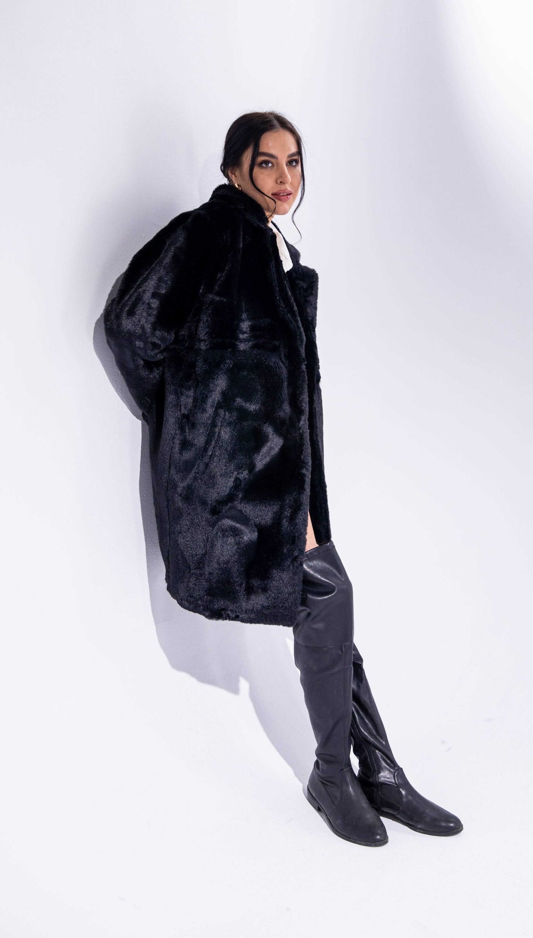 Woman in elegant black faux fur coat and knee-high black boots against white background