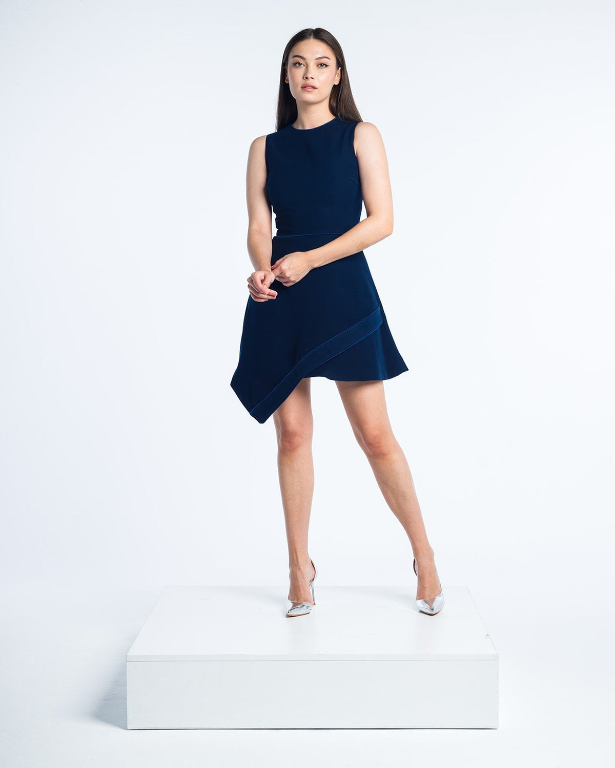 Woman modeling sleeveless navy blue asymmetrical dress and metallic heels on white backdrop