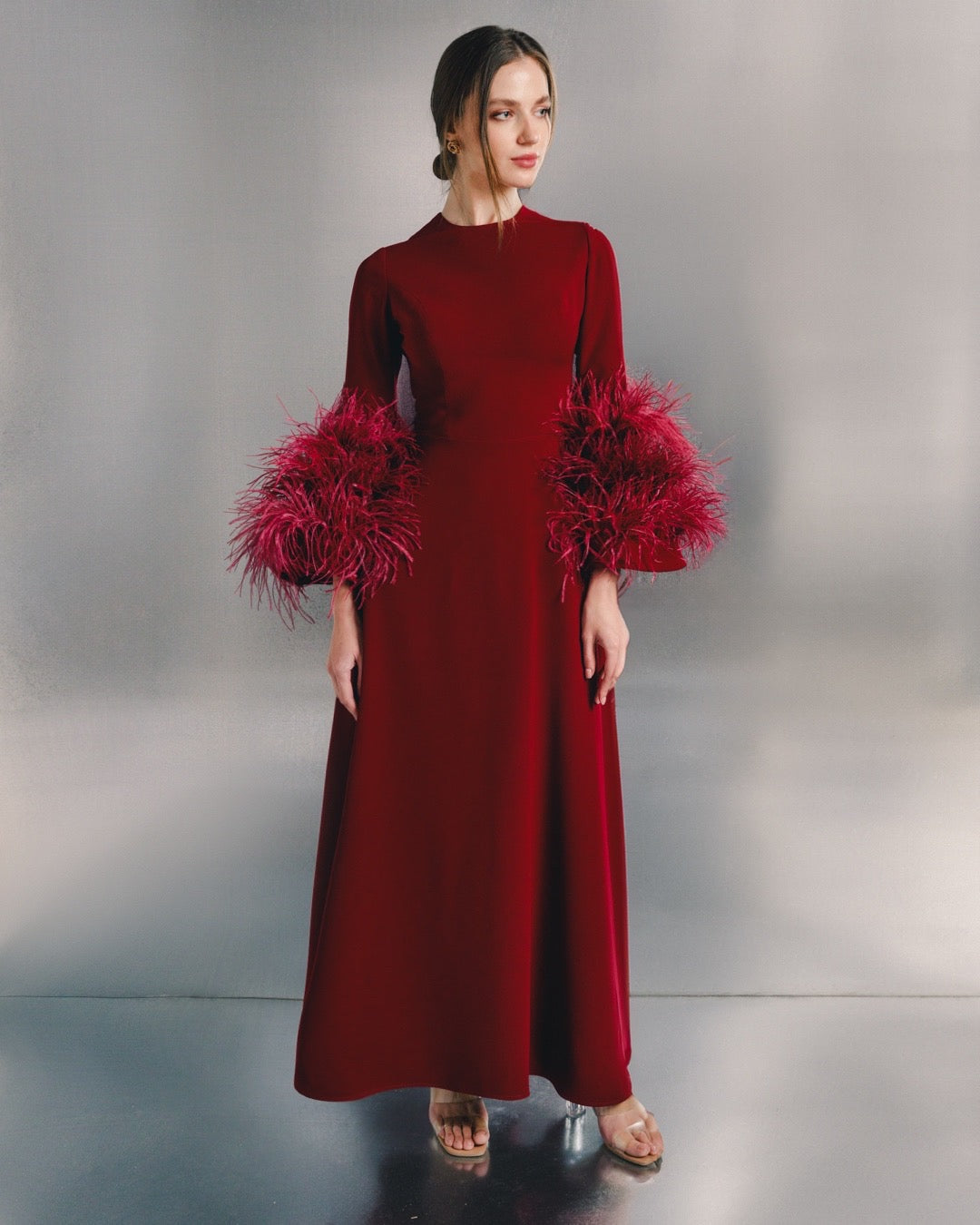 Woman wearing a long red evening dress with dramatic feathered sleeves, standing indoors.