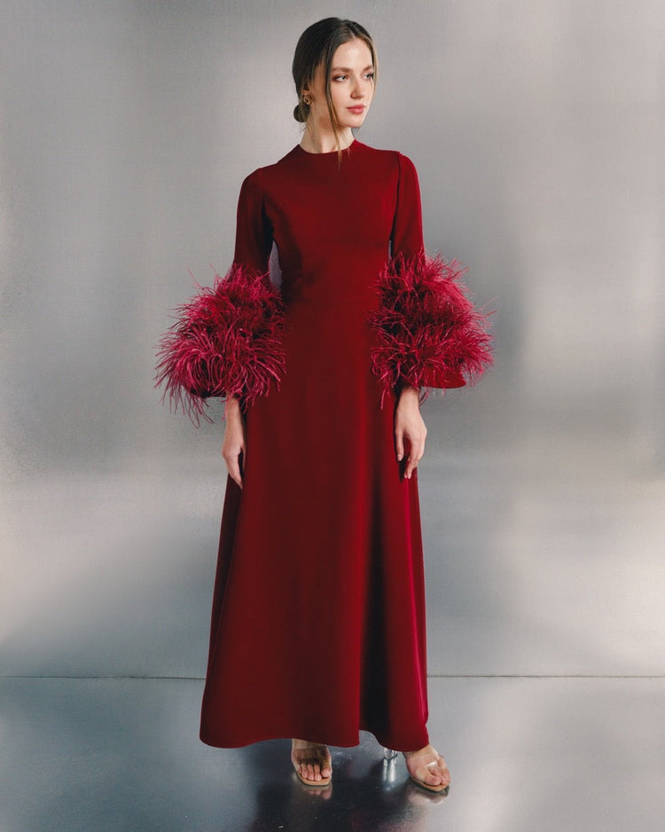 Woman wearing a long red evening dress with dramatic feathered sleeves, standing indoors.