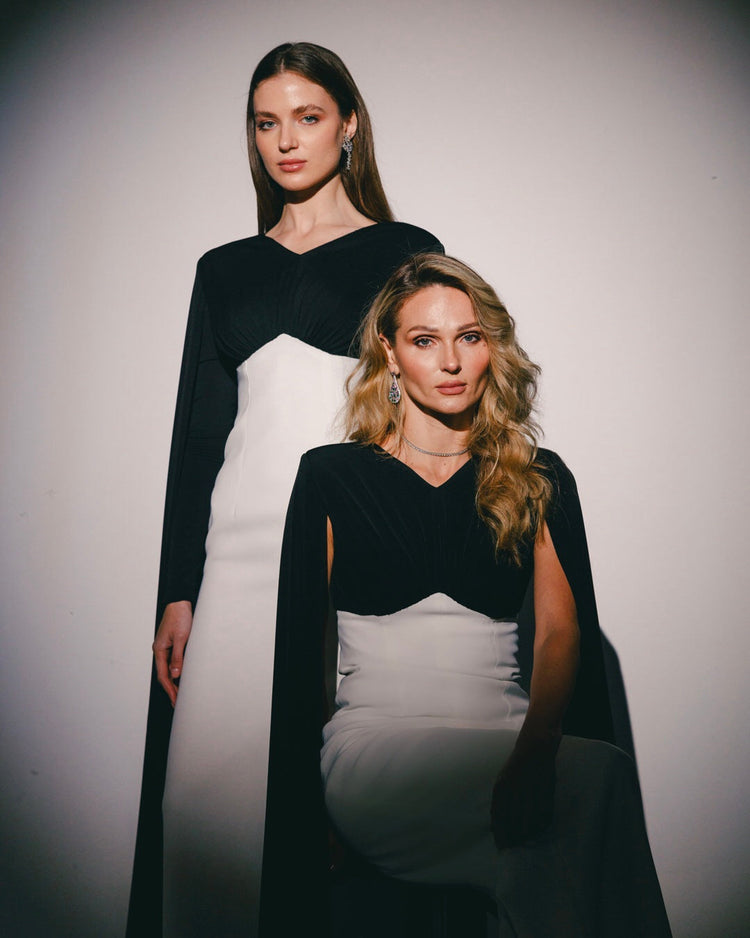 Two women in elegant black and white evening dresses with cape sleeves and statement jewelry