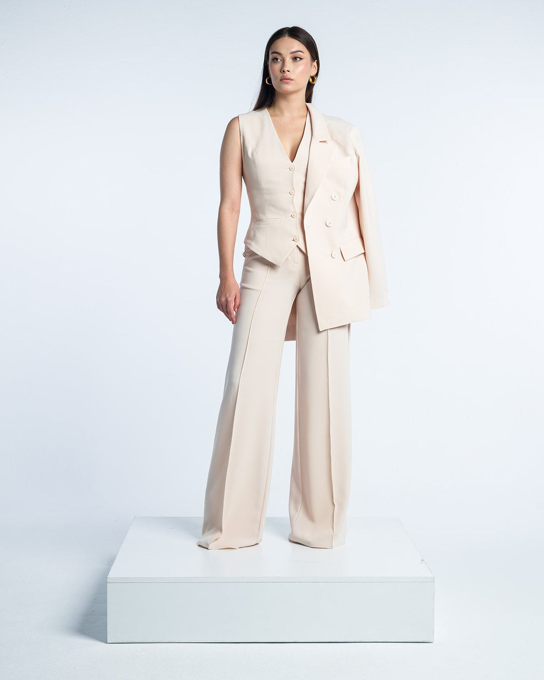 Woman in beige sleeveless suit vest and wide-leg pants, jacket draped over shoulder, studio setting