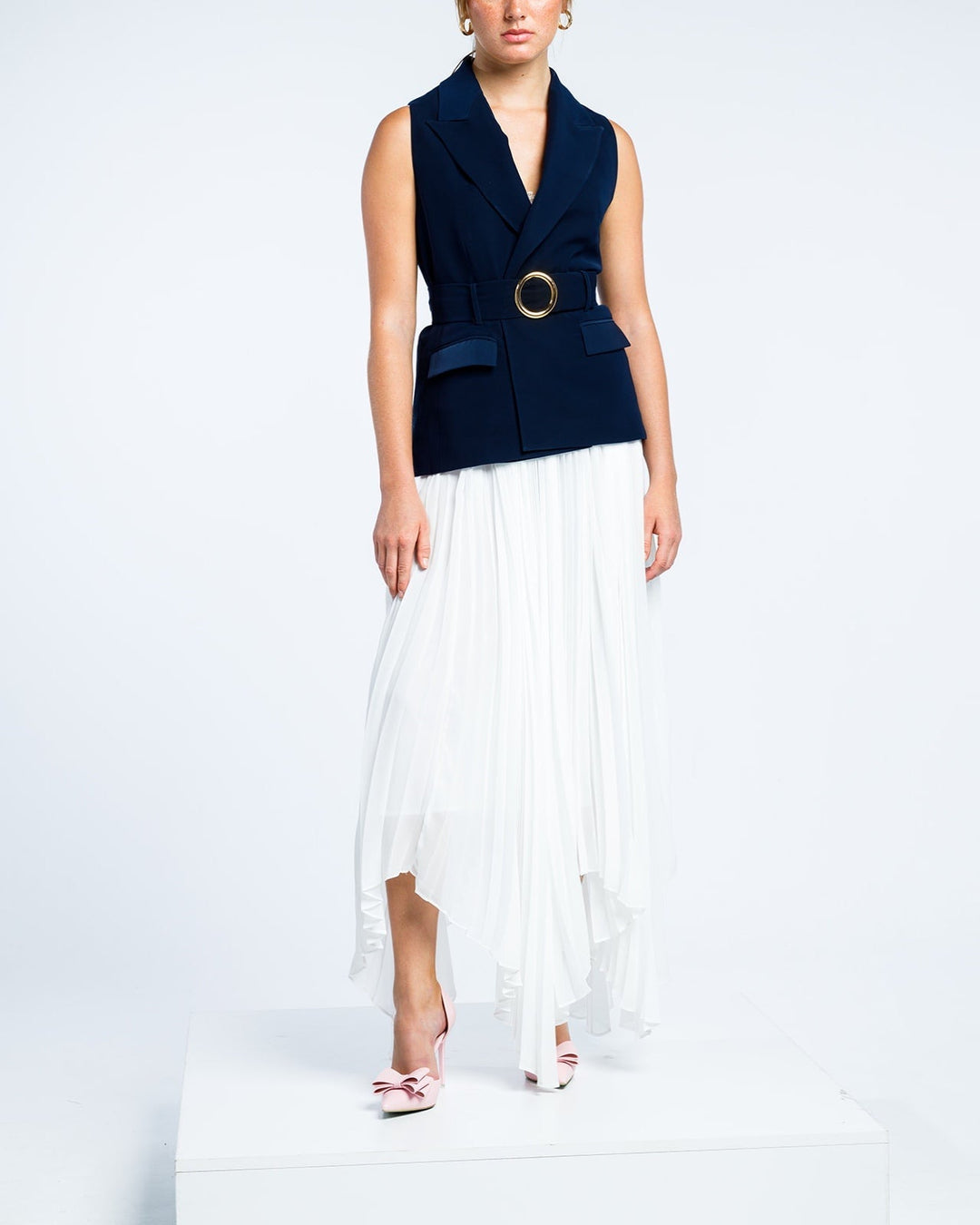 Woman in navy sleeveless blazer with gold buckle belt and white pleated asymmetric skirt