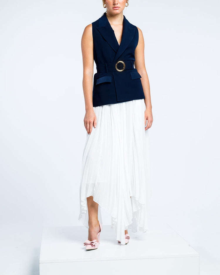 Woman in navy sleeveless blazer with gold buckle belt and white pleated asymmetric skirt