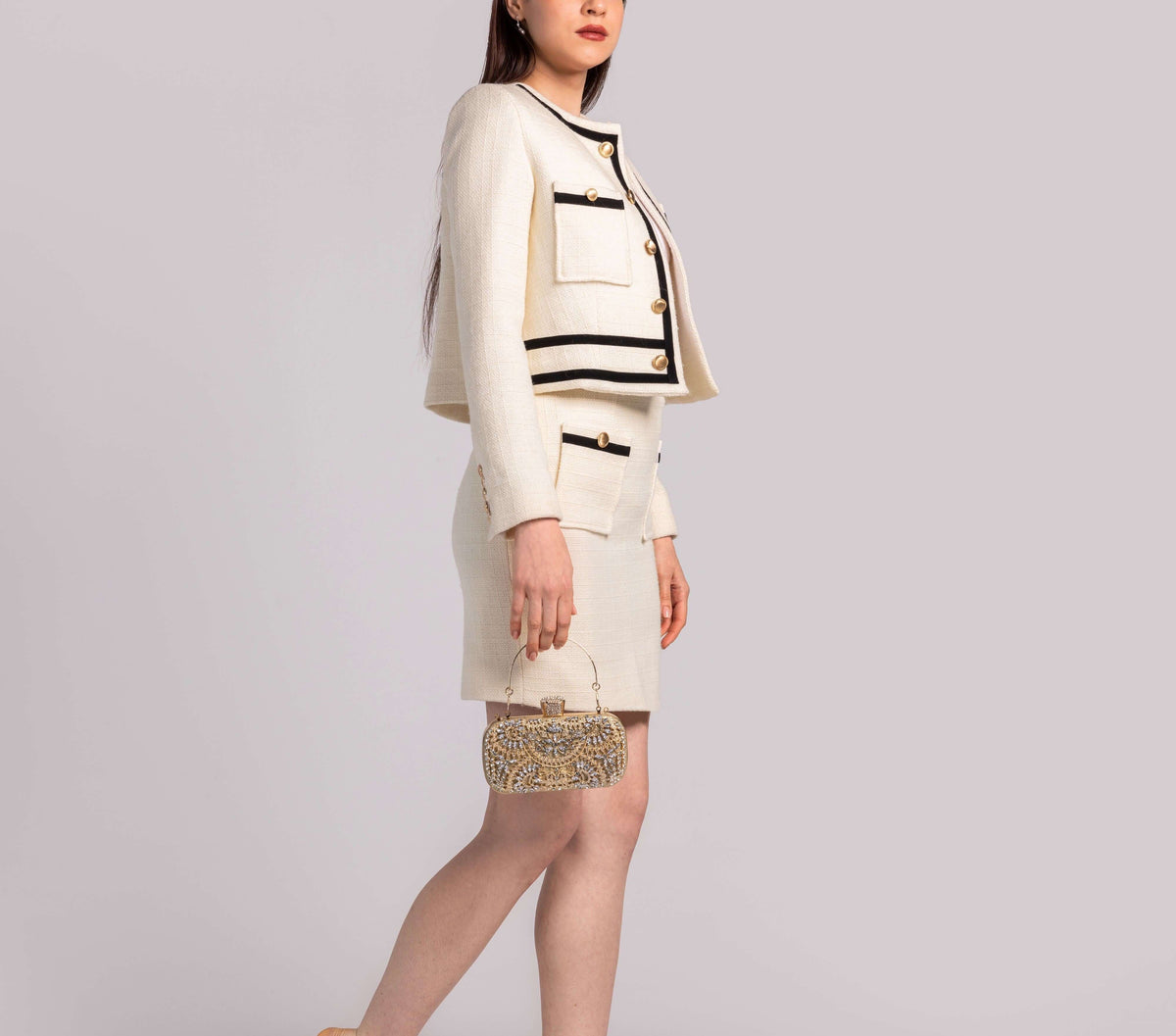 Woman in cream tweed skirt suit with gold buttons, holding embellished clutch bag