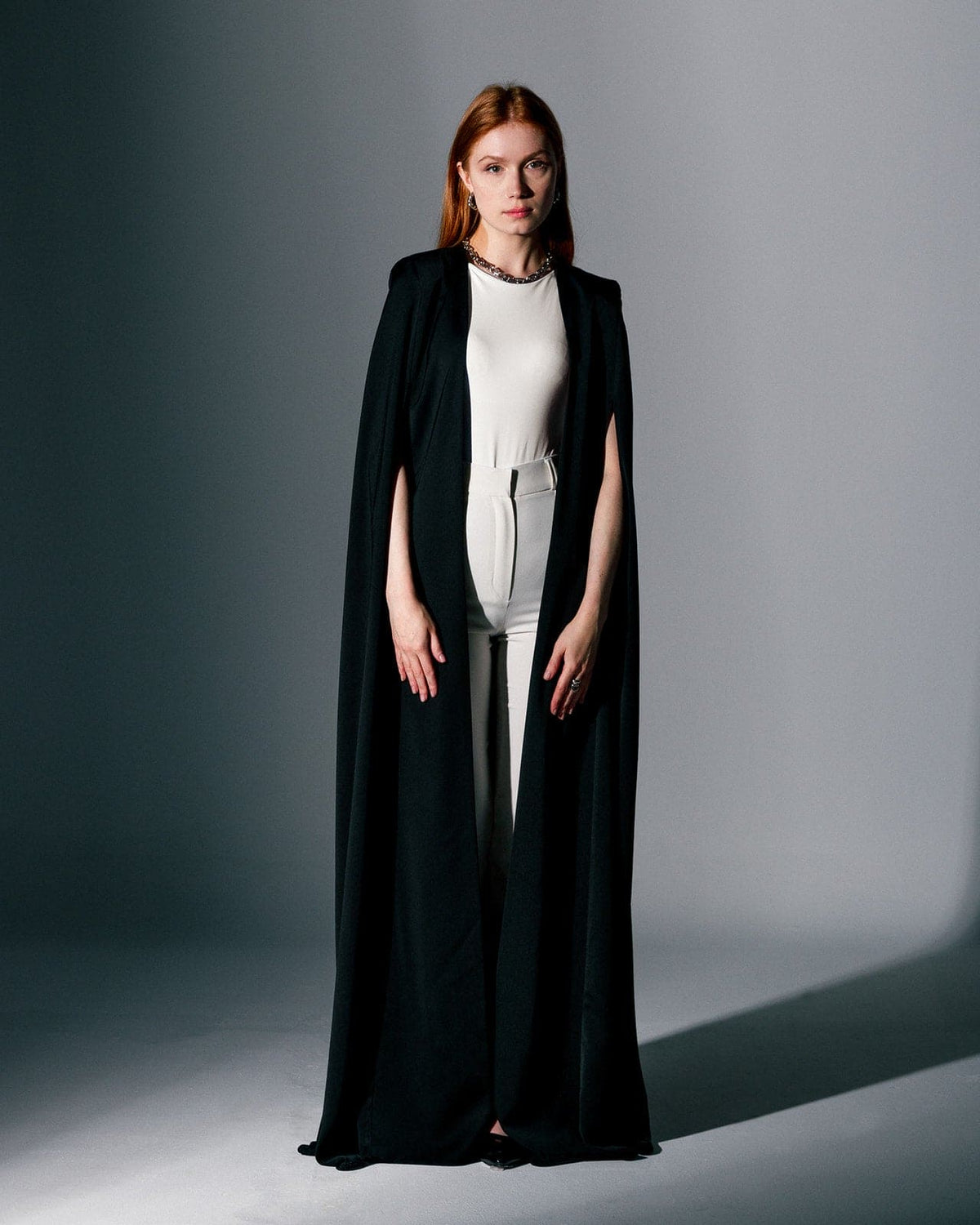 Woman wearing a chic black crepe cape gown over a white jumpsuit in a studio setting