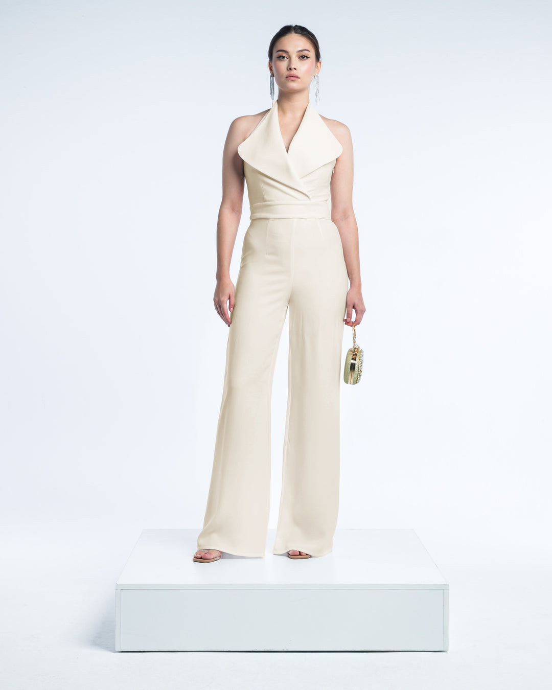 Woman in elegant beige halter jumpsuit on platform, holding gold textured clutch