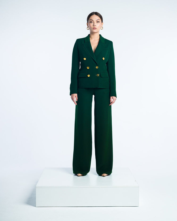 Woman in dark green double-breasted suit with gold buttons and wide-leg pants on white platform