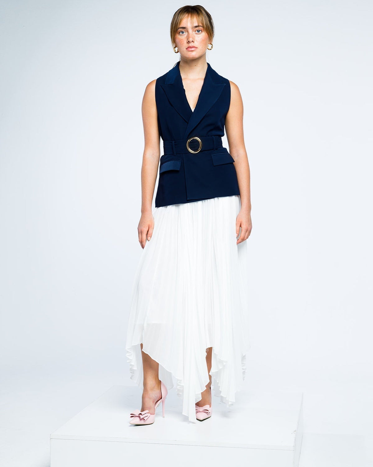 Woman in navy blue sleeveless blazer with gold belt, white pleated skirt, pink heels