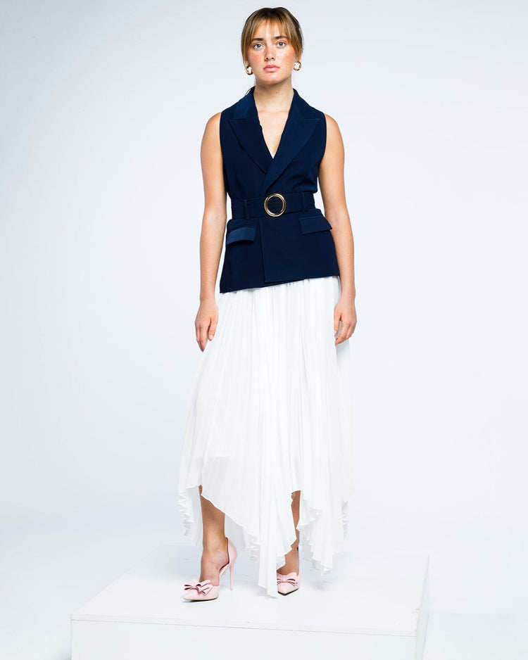 Woman in navy blue sleeveless blazer with gold belt, white pleated skirt, pink heels