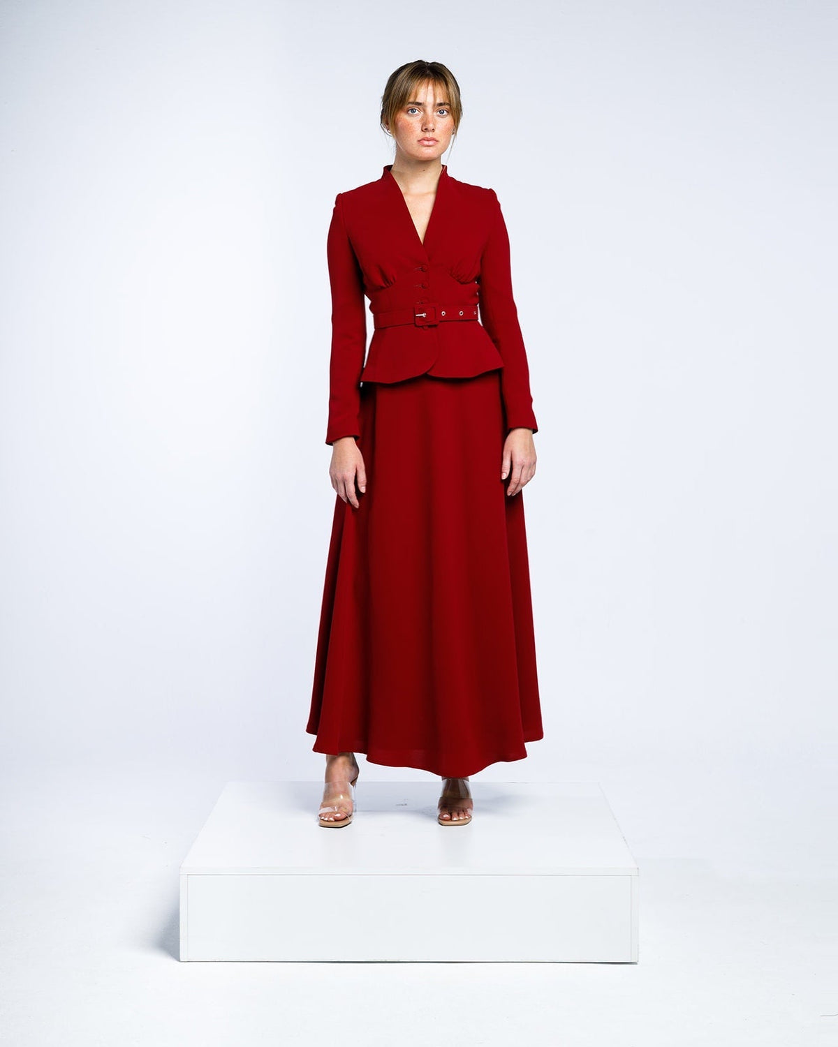 Woman in an elegant long-sleeve red belted maxi dress with a v-neck standing on a white platform