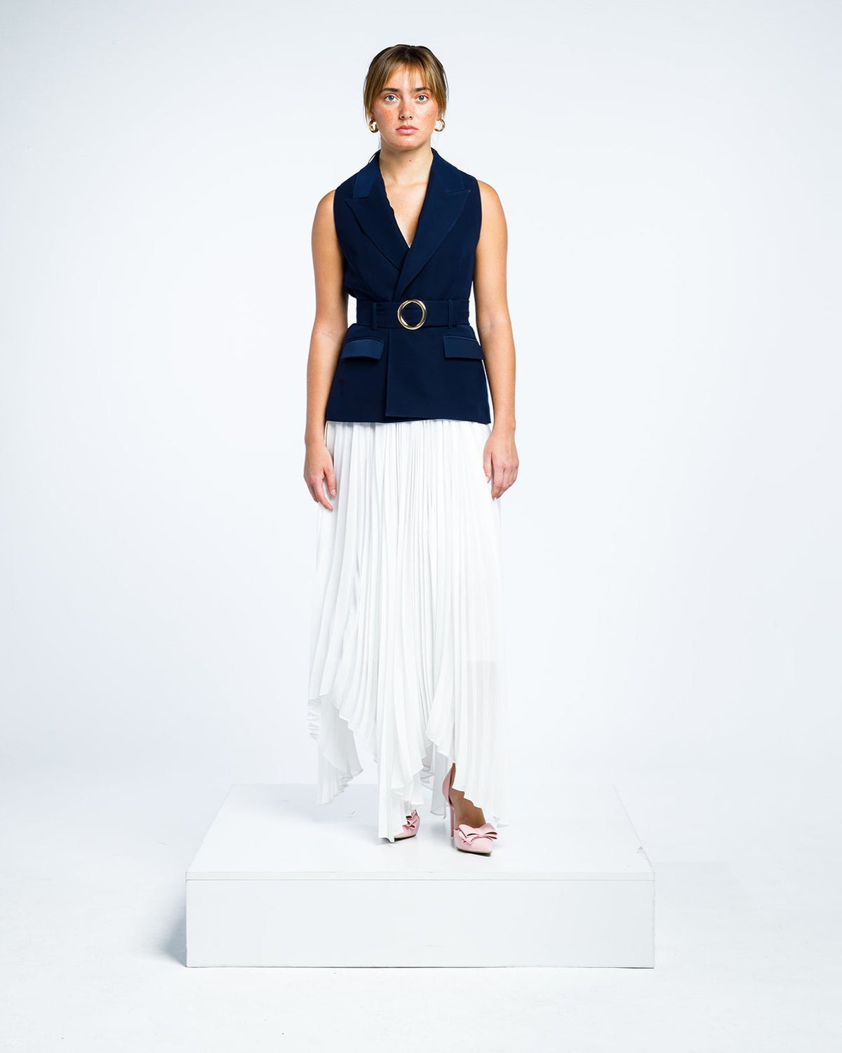 Woman modeling a sleeveless navy blue belted blazer with white pleated maxi skirt and pink heels
