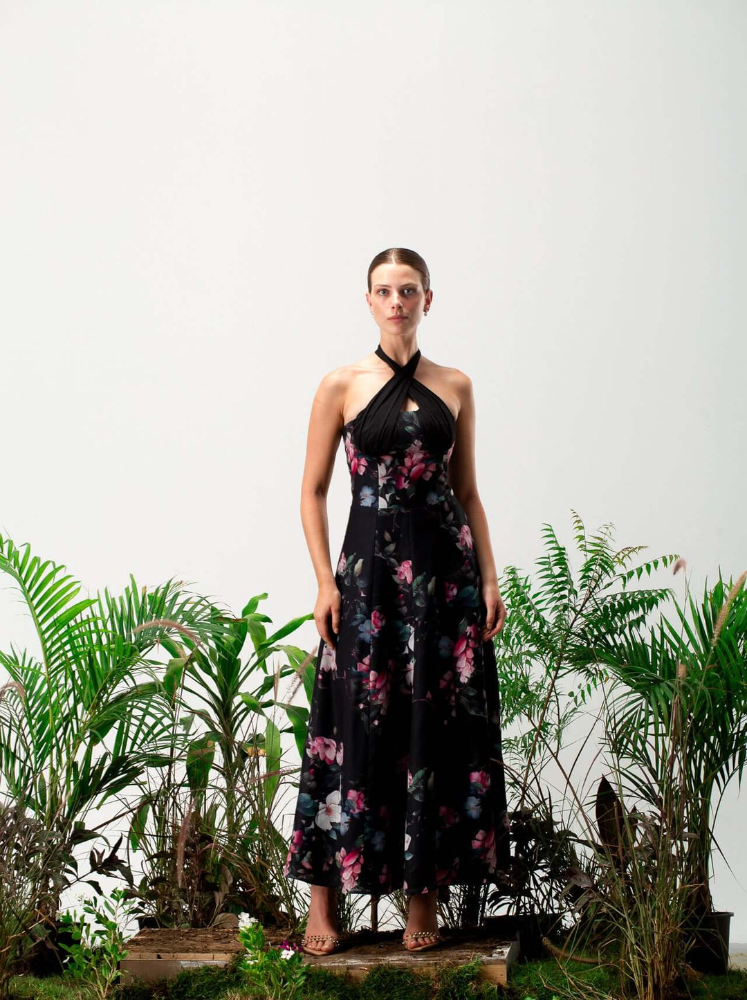 Woman in a black floral halter maxi dress standing among green plants, elegant style