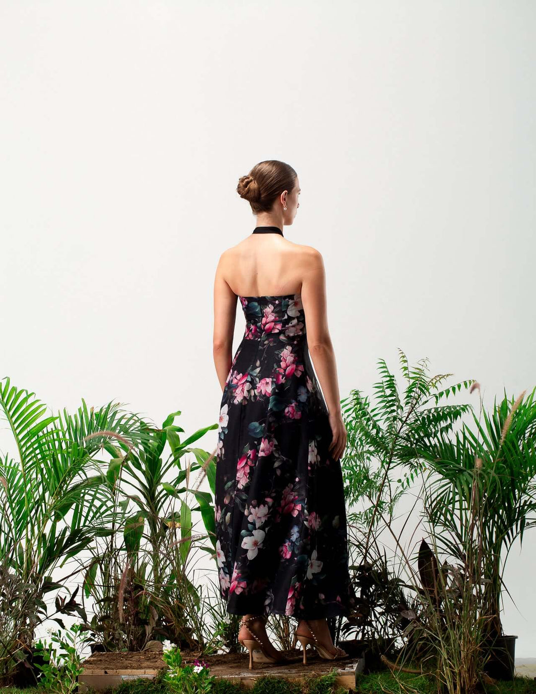 Woman in a strapless black floral dress with halter neck, standing among green plants