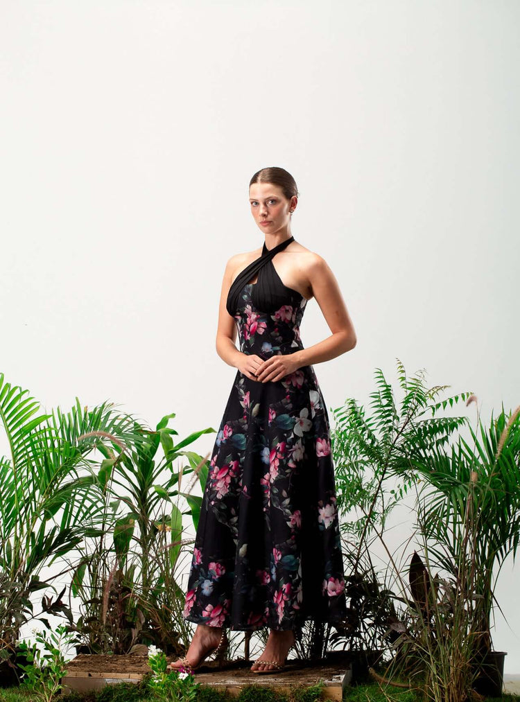 Woman in a black floral halter-neck dress posing among green plants against a white background