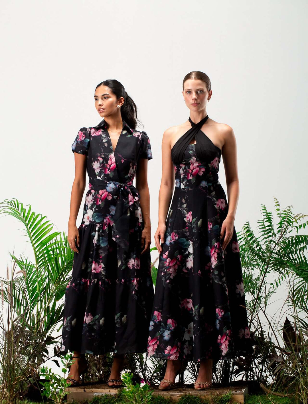 Two women in elegant black floral maxi dresses standing among green plants
