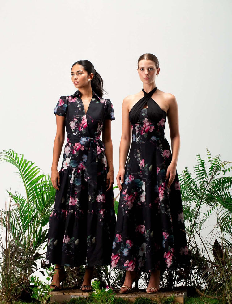 Two women in elegant black floral maxi dresses standing among green plants