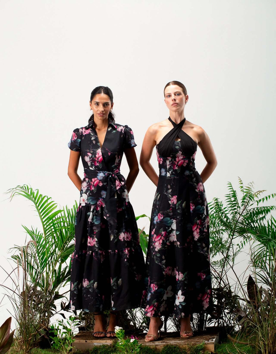 Two women wearing elegant black floral maxi dresses standing among green plants