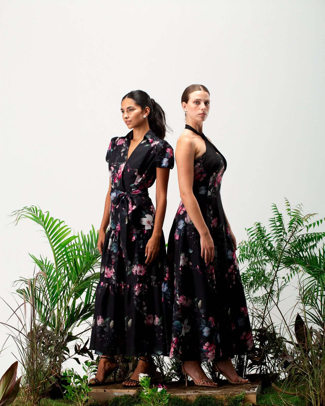 Two women in elegant black floral maxi dresses posing among green plants