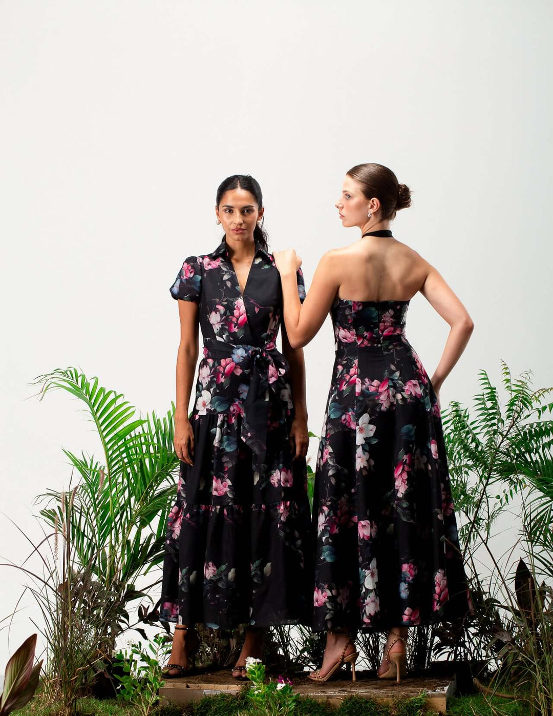 Two women modeling elegant black floral maxi dresses in a studio with green plants