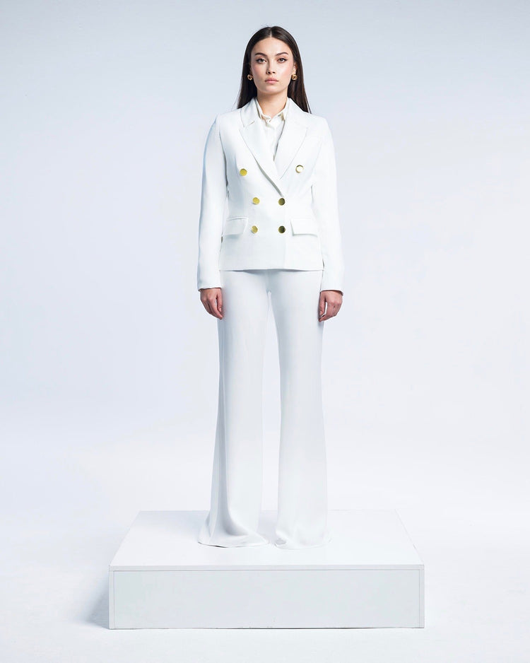 Woman in elegant white suit with gold buttons and flared pants, standing on white platform