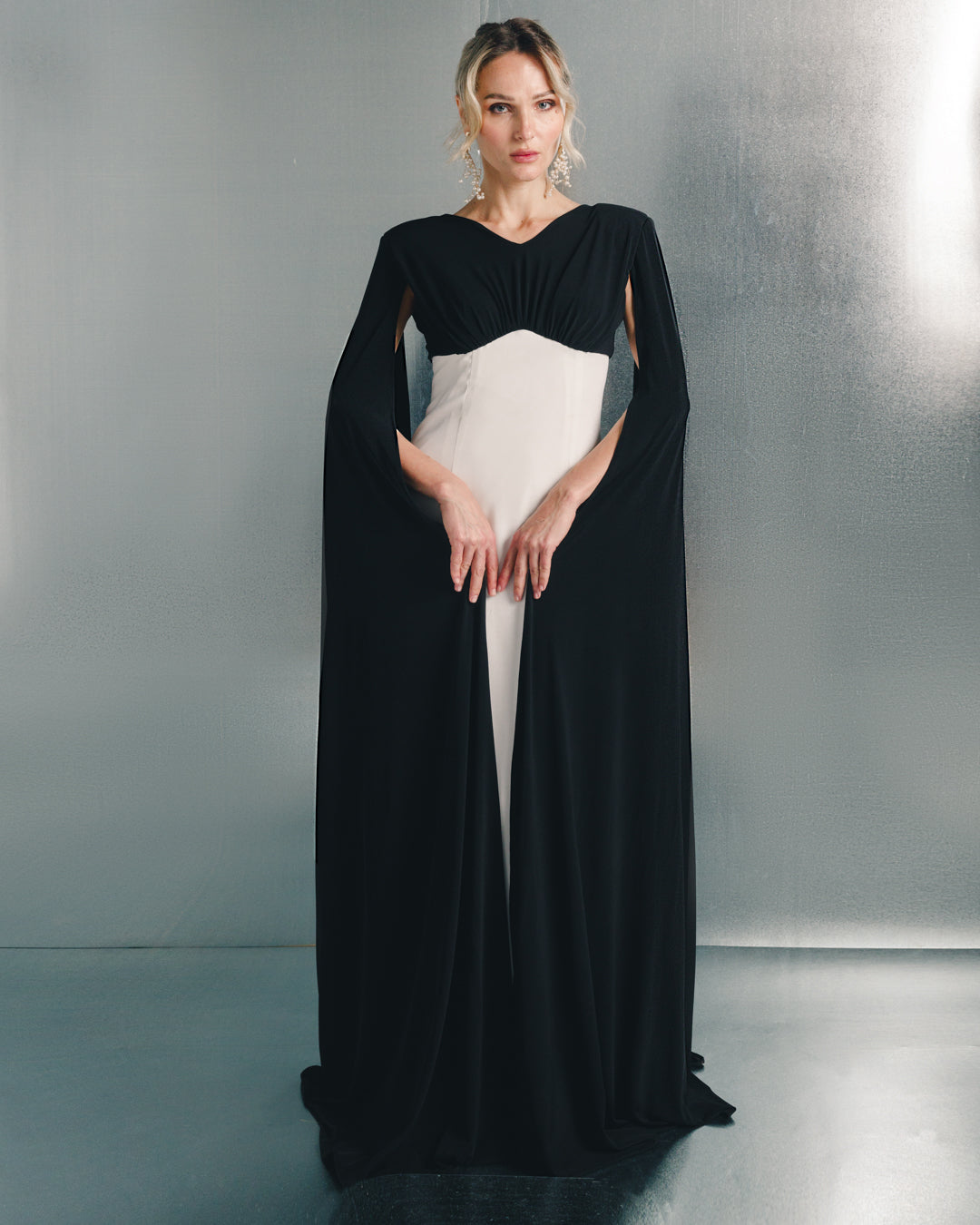 Woman in a black and white elegant floor-length gown with cape sleeves, standing indoors.