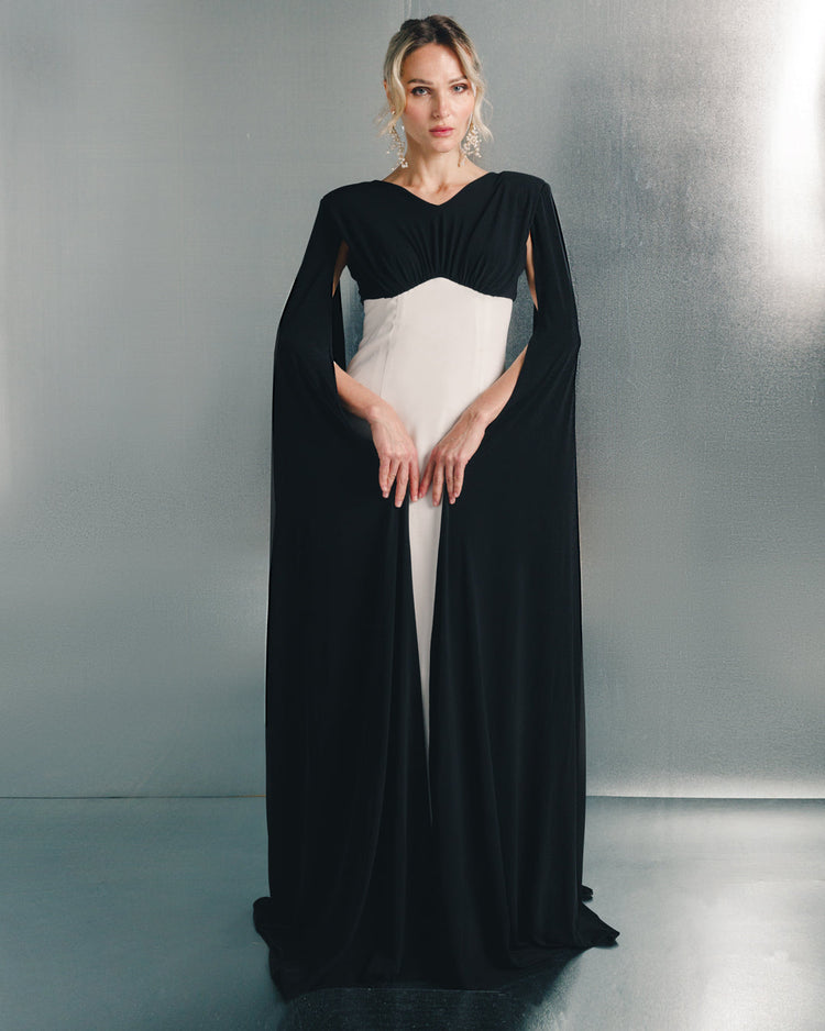 Woman in a black and white elegant floor-length gown with cape sleeves, standing indoors.