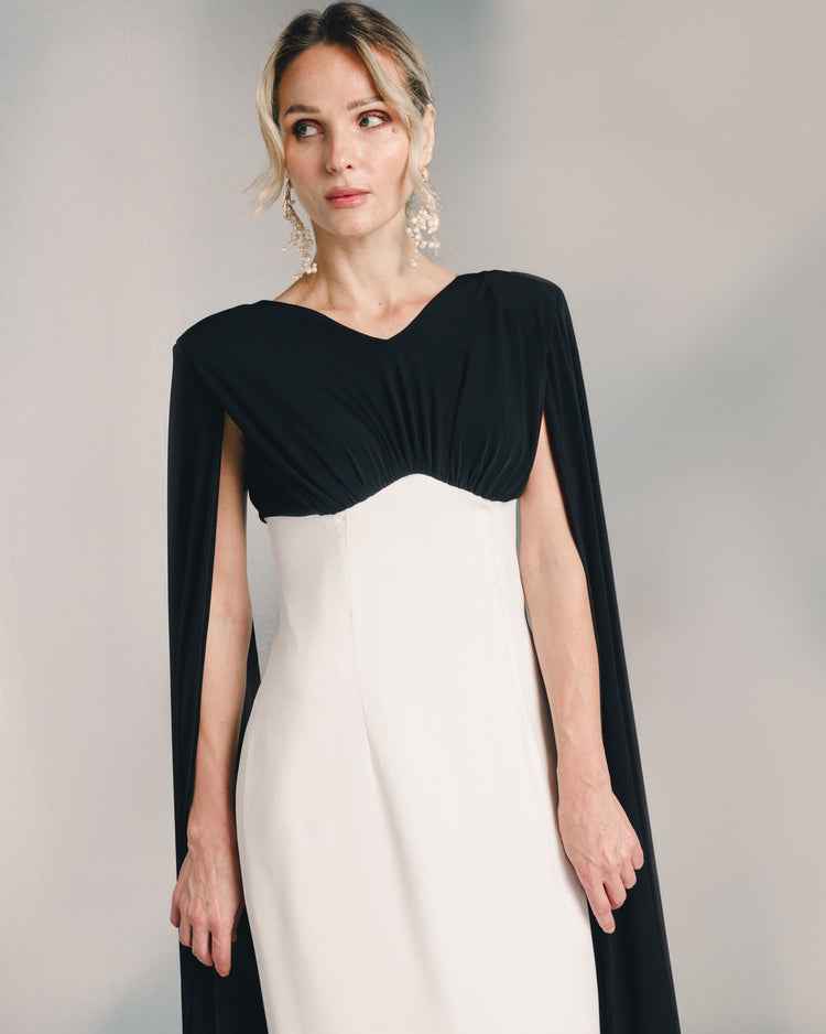 Woman wearing elegant black and white cape dress with statement earrings against neutral background.