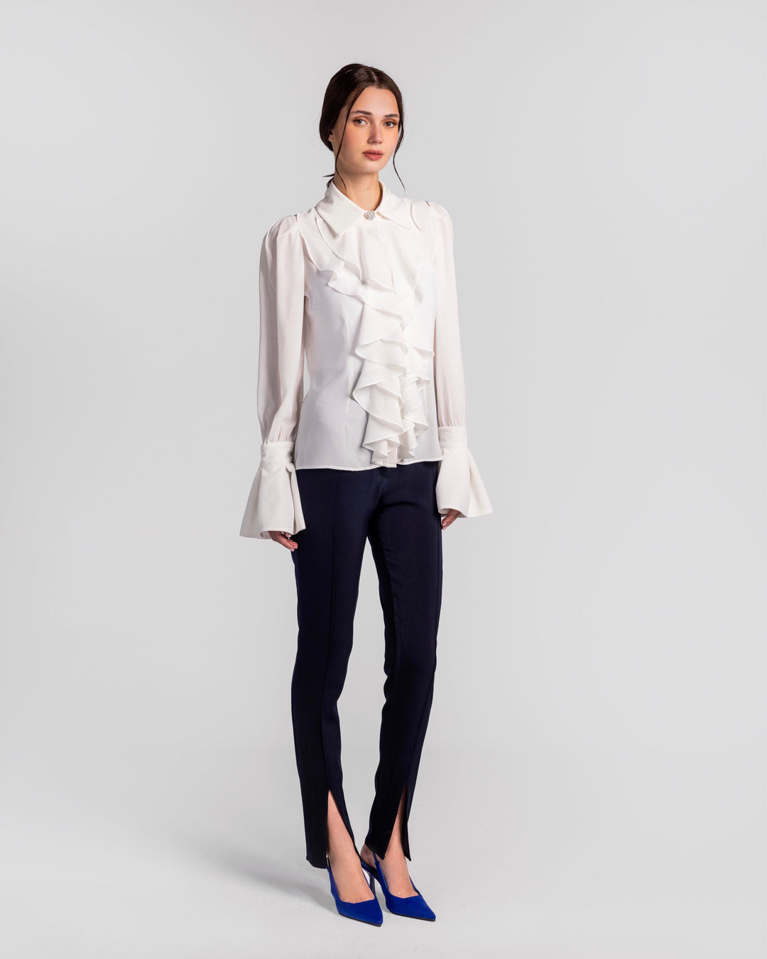 Woman wearing white ruffled blouse, slim navy pants with front slits, blue heels; studio backdrop