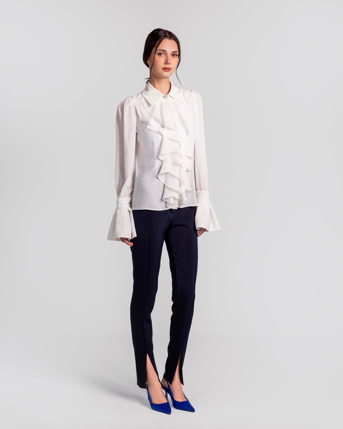 Woman wearing white ruffled blouse, slim navy pants with front slits, blue heels; studio backdrop