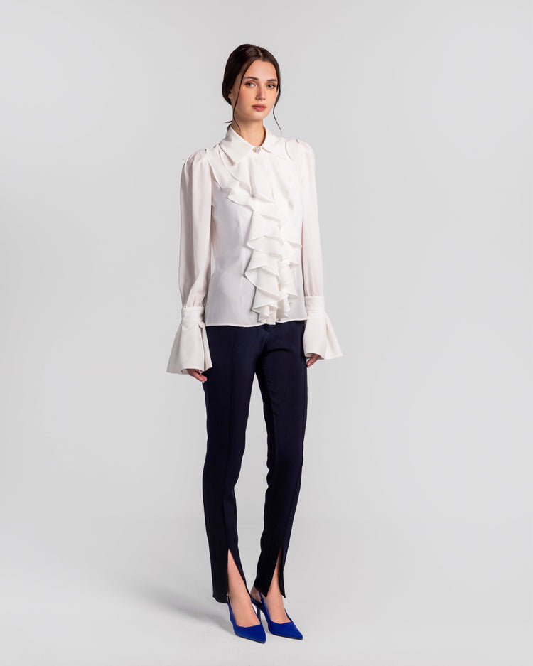 Woman wearing white ruffled blouse, slim navy pants with front slits, blue heels; studio backdrop
