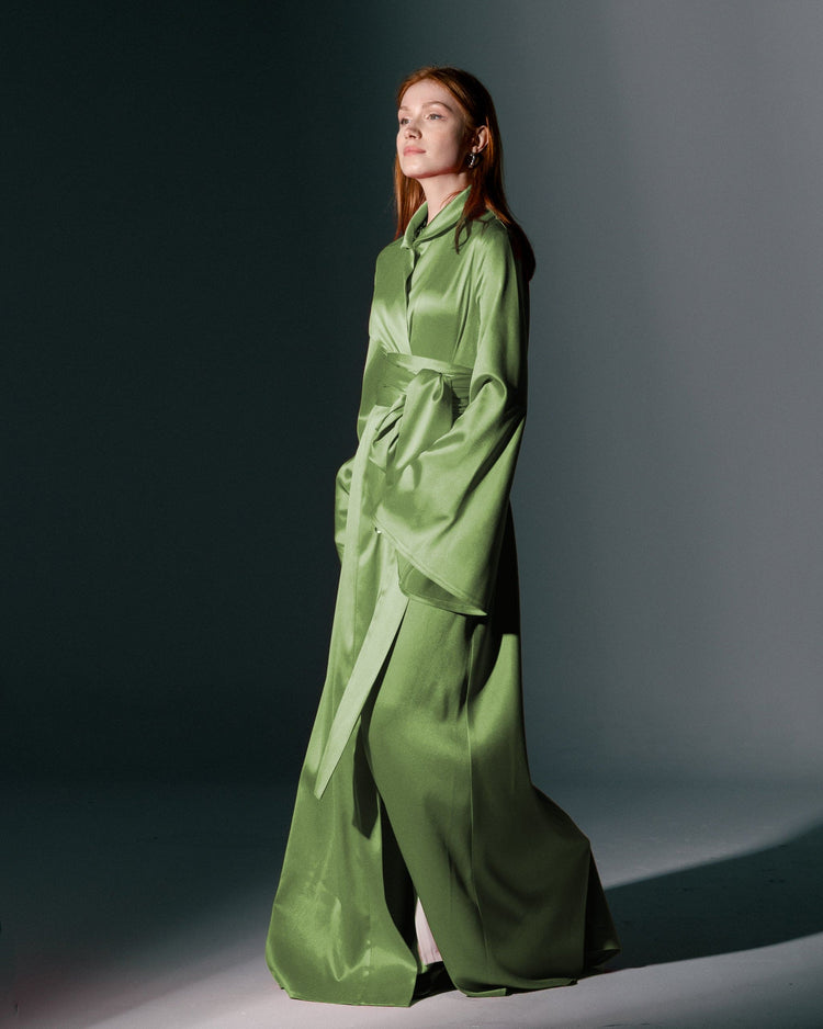 Woman in elegant long green satin dress with wide sleeves and belt on studio background