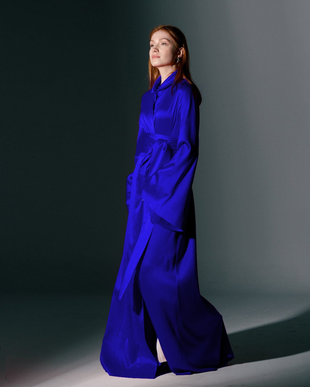 Woman in a vibrant blue satin maxi dress with wide sleeves and sash belt, studio lighting