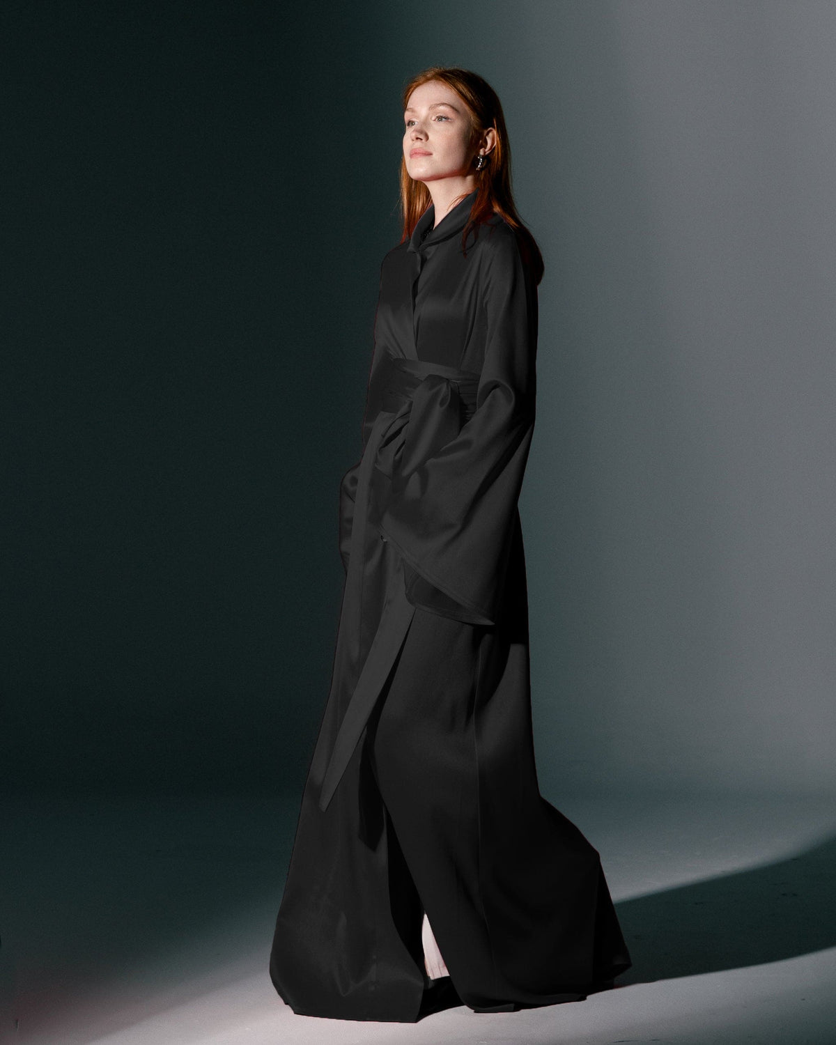Woman in elegant black long-sleeve dress with belt, standing in studio spotlight