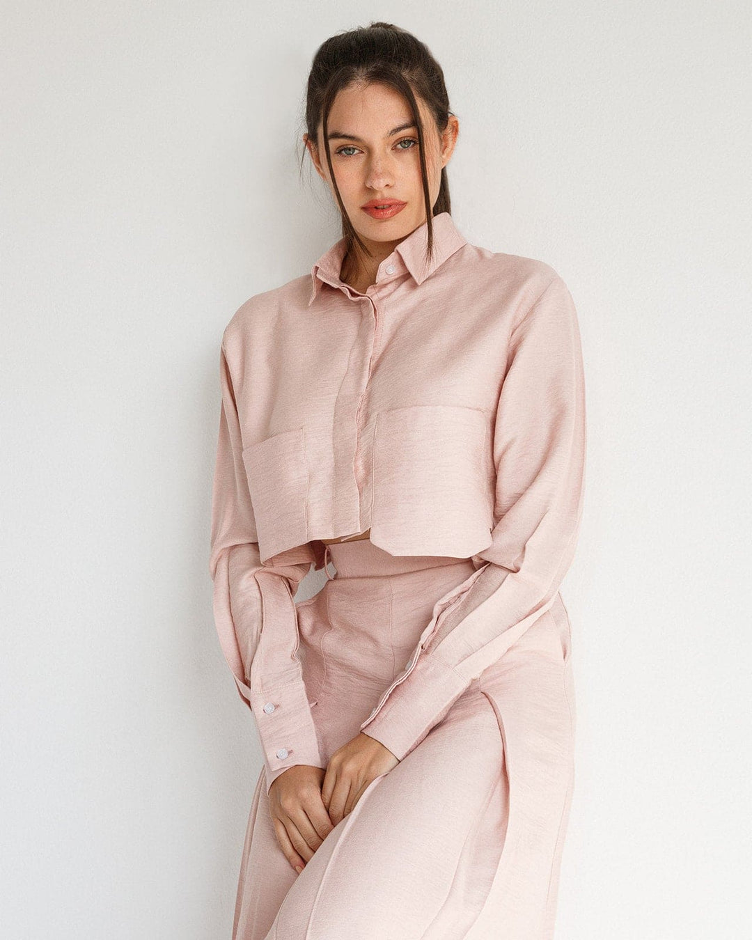 Woman wearing a light pink cropped shirt and matching high-waist skirt against a white wall