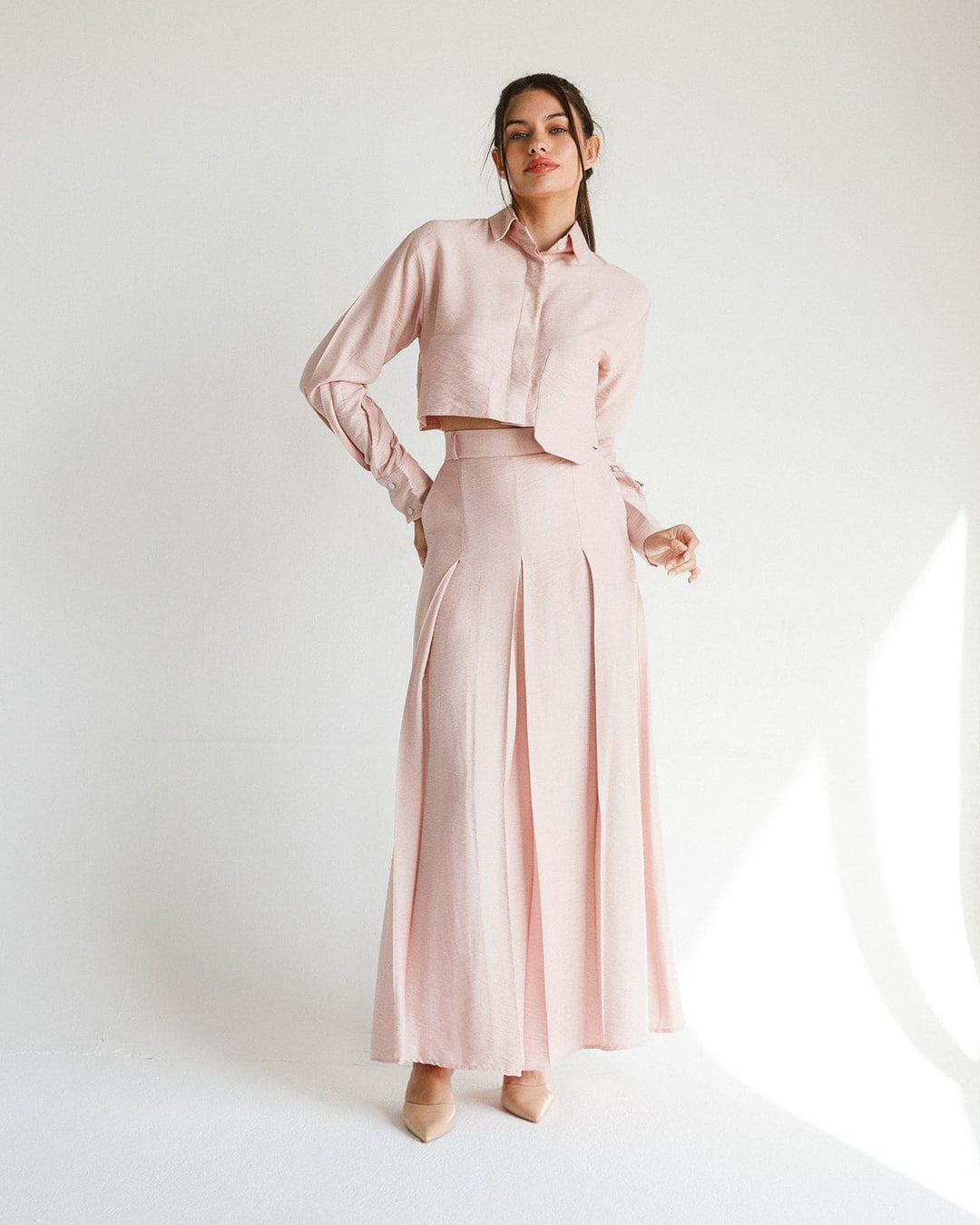 Woman wearing light pink two-piece set with cropped shirt and long pleated skirt in minimal studio