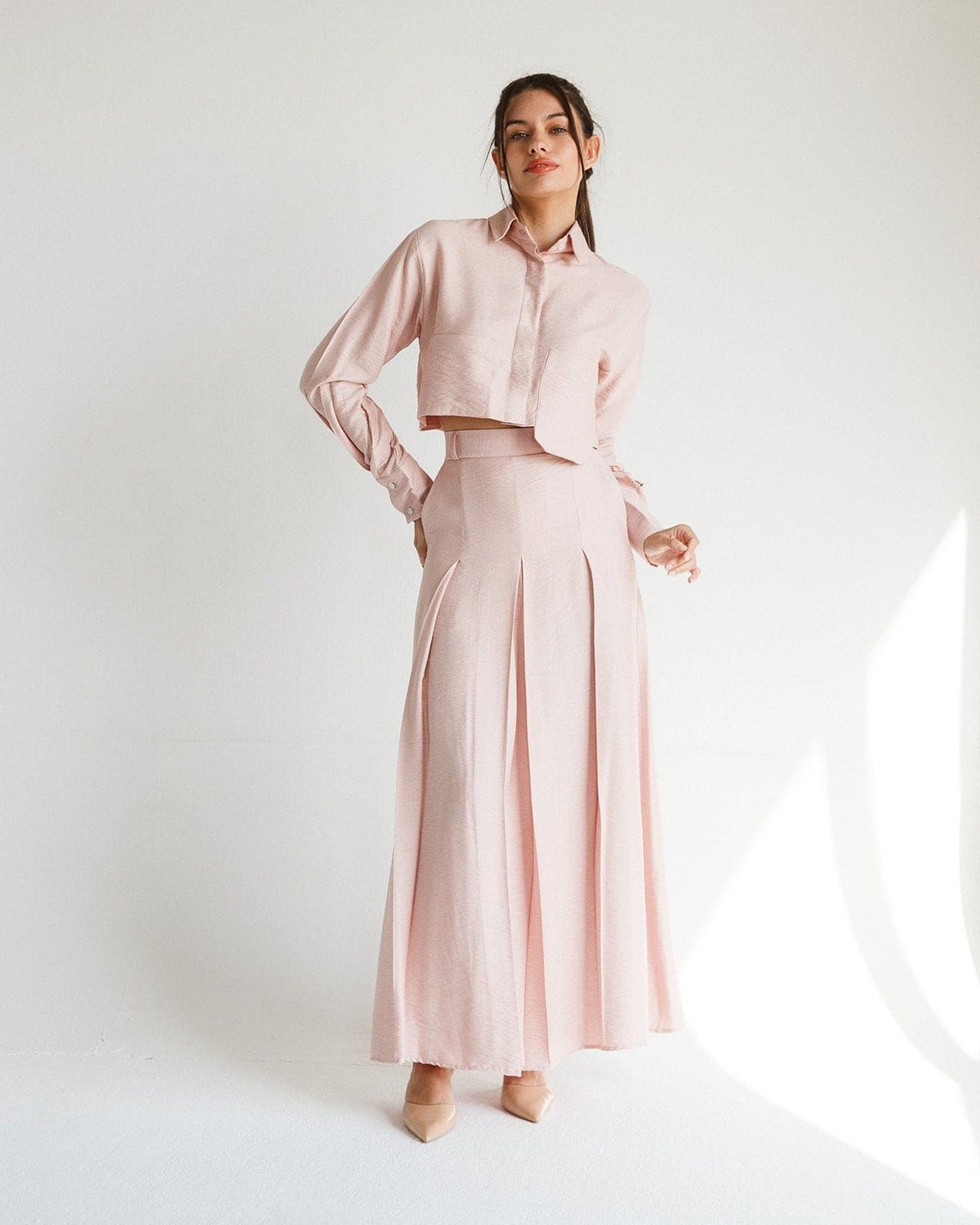 Woman wearing light pink two-piece set with cropped shirt and long pleated skirt in minimal studio