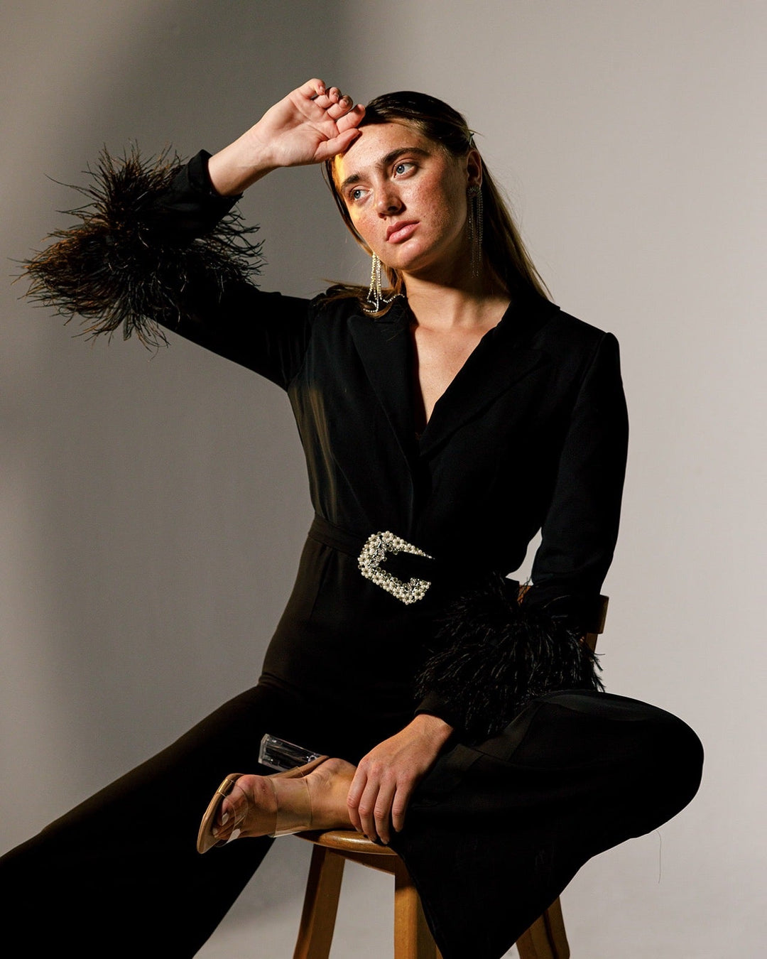 Woman in elegant black jumpsuit with feather cuffs and crystal belt, seated on a stool